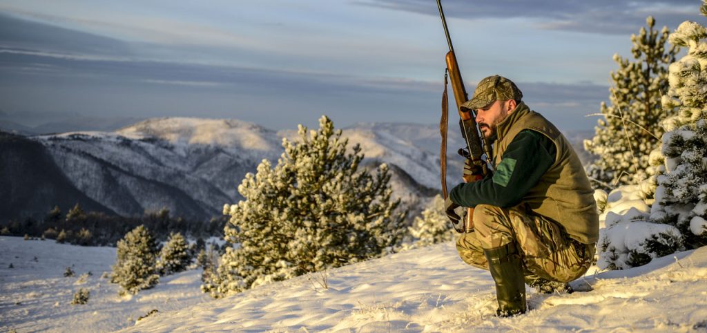 Hunting guide with a rifle crouched on top of a snowy mountain.